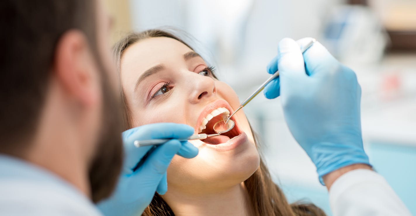 Dentist examining patient teeth with a mouth mirror and dental excavator. Close-up view on the woman's face