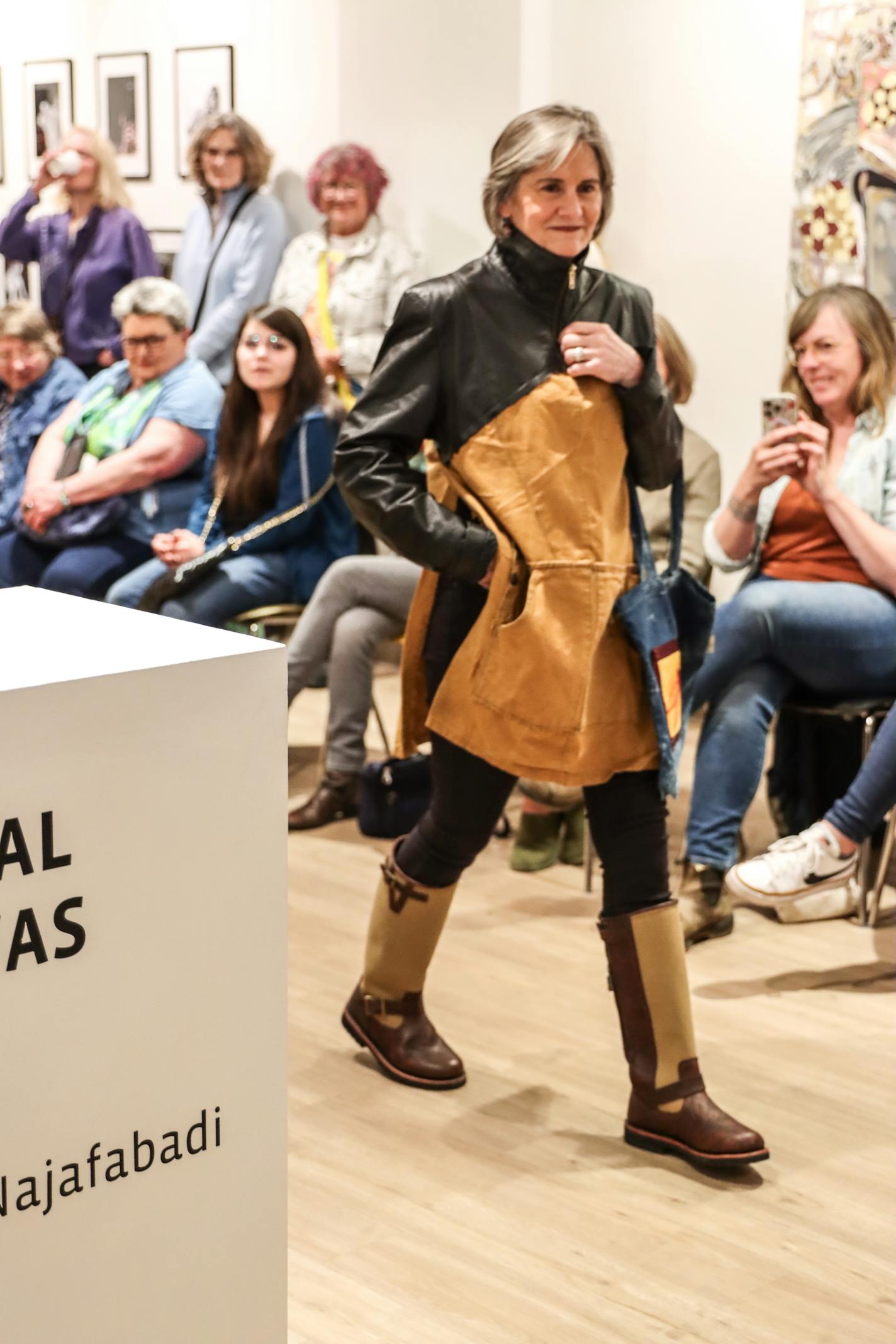 A woman with gray hair smiles while wearing a two-toned leather jacket and tall boots in an art gallery. Many onlookers are seated along her walkway.