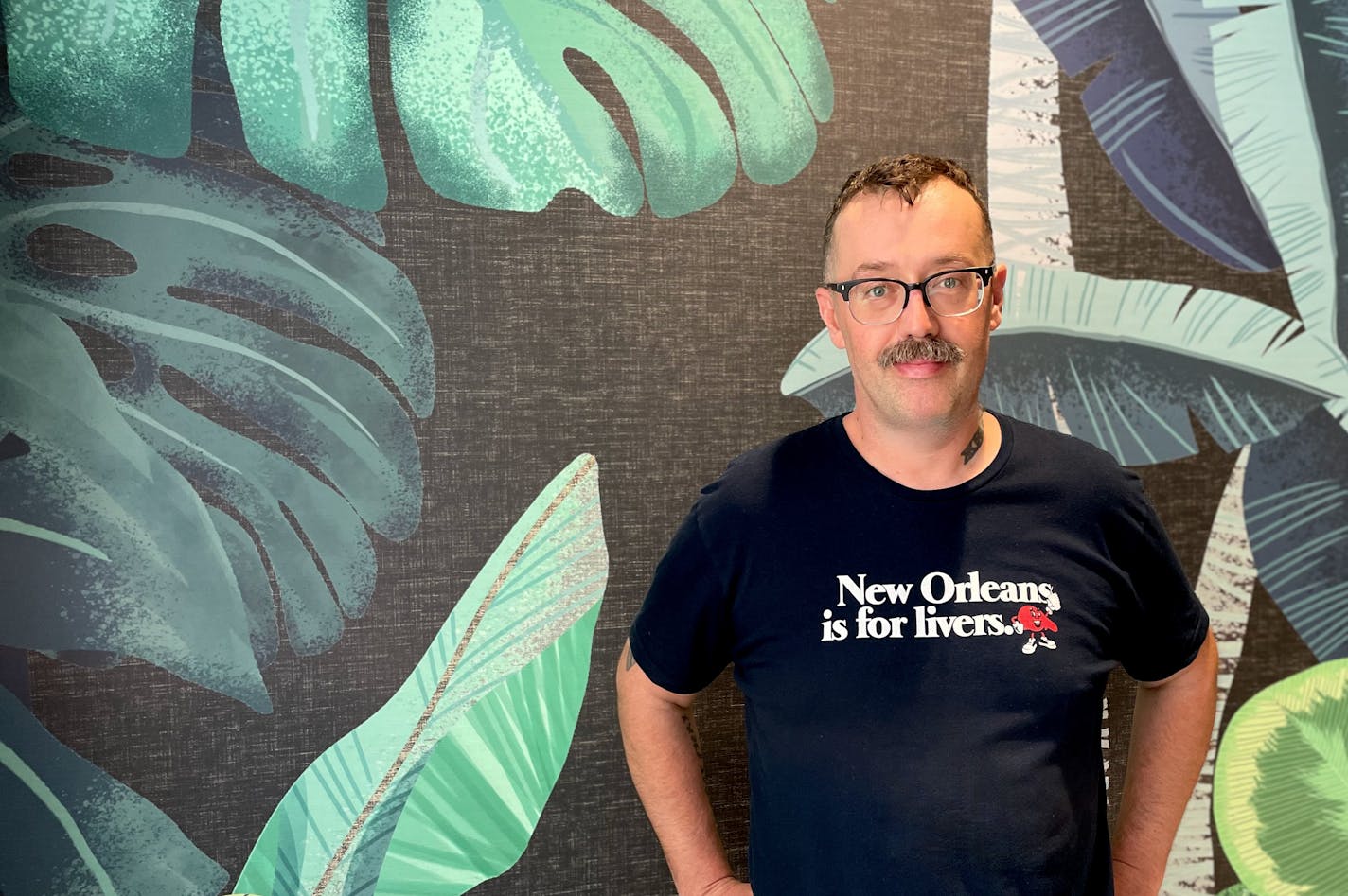 Serge Kogan in front of a tropical-leaf wallpapered wall wearing a T-shirt that says "New Orleans is for livers."