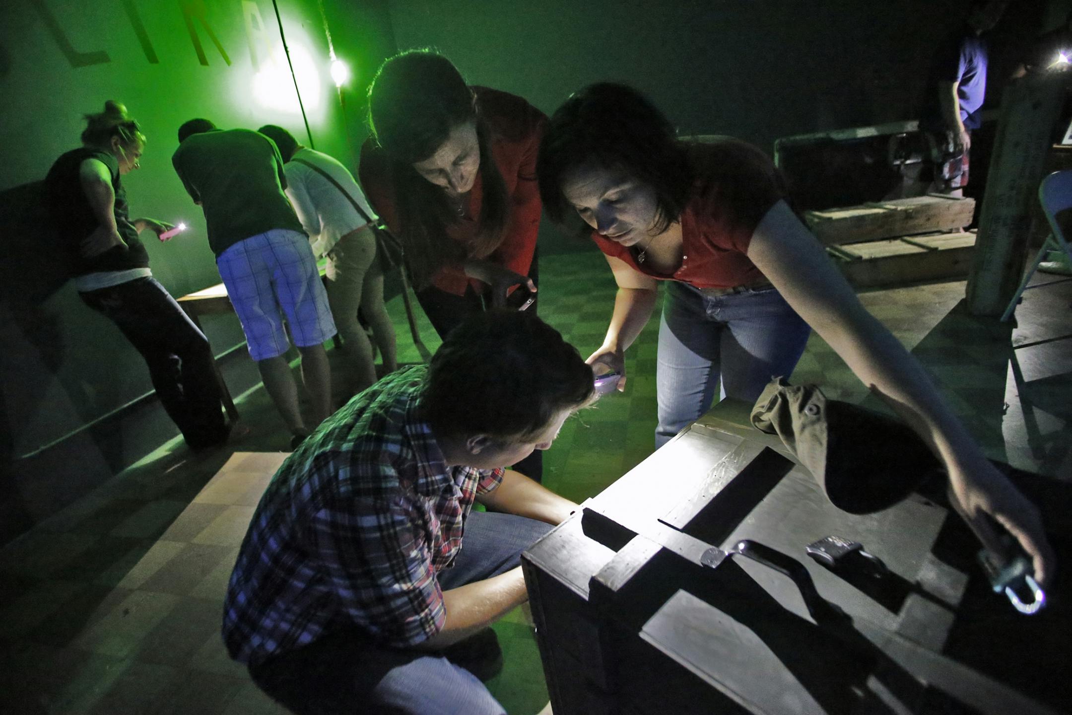 A group works on the various clues presented to them in an effort to find passage out of the riddle room. Some attempt to unlock a chest in foreground while others seek clues from a map at left.