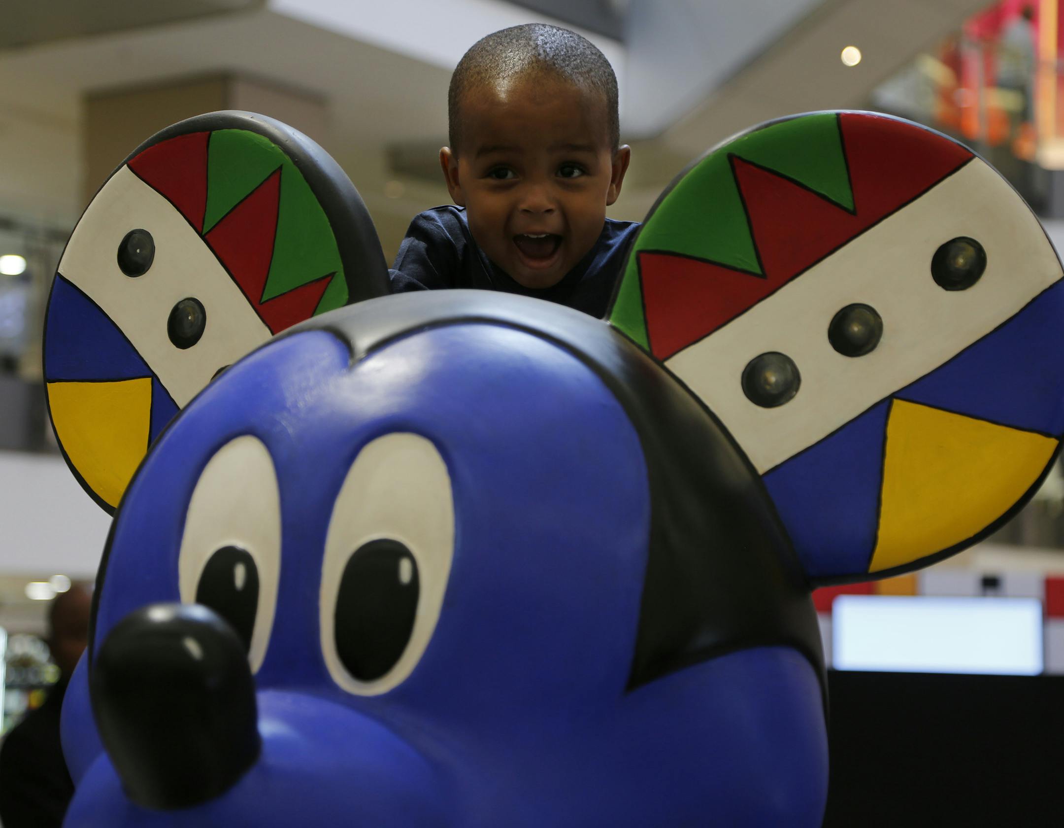A child looks over the head of a Mickey Mouse statue at an exhibition at Sandton City in Johannesburg, Sunday Sept. 30, 2018. For the famed cartoon's 90th birthday and at Disney's invitation, 10 South African artists each painted and adorned a statue of the cartoon character, using colors, patterns and imagery that reflect the continent's cultural diversity and giving him more wardrobe options than his usual white gloves, red pants and yellow shoes. (AP Photo/Denis Farrell) ORG XMIT: XDF101