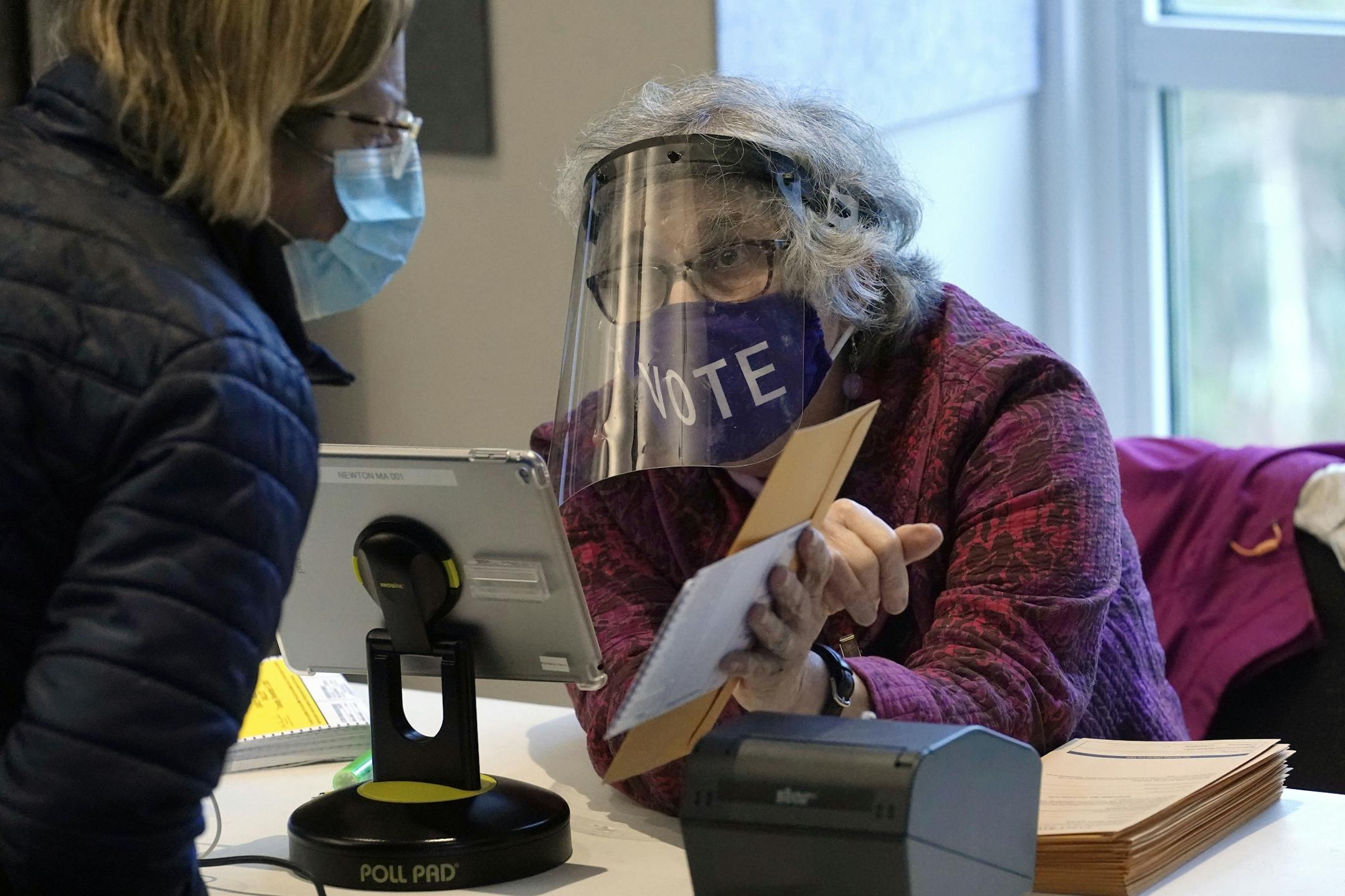 In this Oct. 28, 2020, photo, poll worker Alice Machinist, of Newton, Mass., right, wears a mask and shield out of concern for the coronavirus while assisting a voter, left, with a ballot during early in-person general election voting at the Newton Free Library, in Newton, Mass.