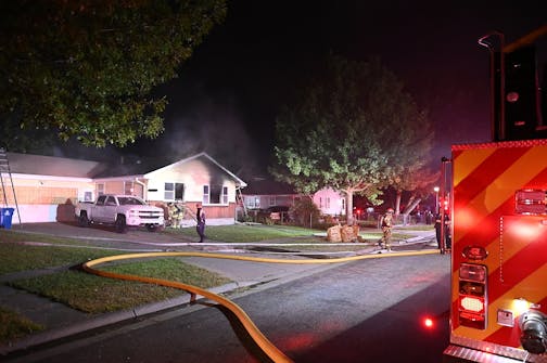 Small yellow and white house in background with smoke coming from window, with firetruck at right foreground and hoses on the lawn and firefighters working in early morning darkness.