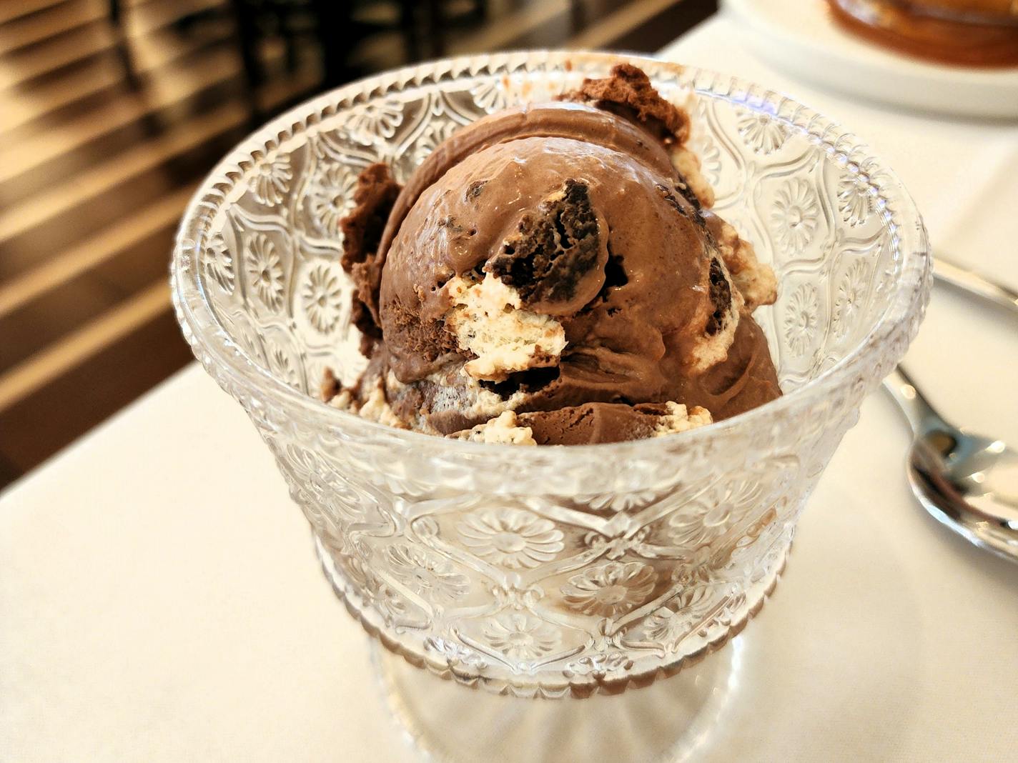 An elegant etched glass coupe sits on a white tablecloth. Inside is a dark brown scoop of ice cream studded with black chocolate brownies and white marshmallow bits.