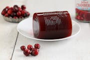 A close up horizontal photograph of some commercially canned cranberry sauce on a small white plate surrounded by fresh cranberries and a can.