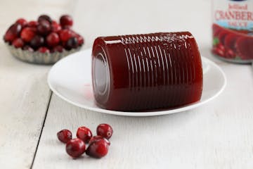 A close up horizontal photograph of some commercially canned cranberry sauce on a small white plate surrounded by fresh cranberries and a can.