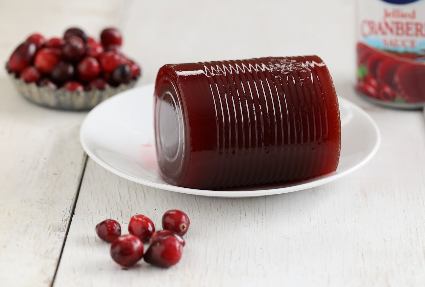 A close up horizontal photograph of some commercially canned cranberry sauce on a small white plate surrounded by fresh cranberries and a can.