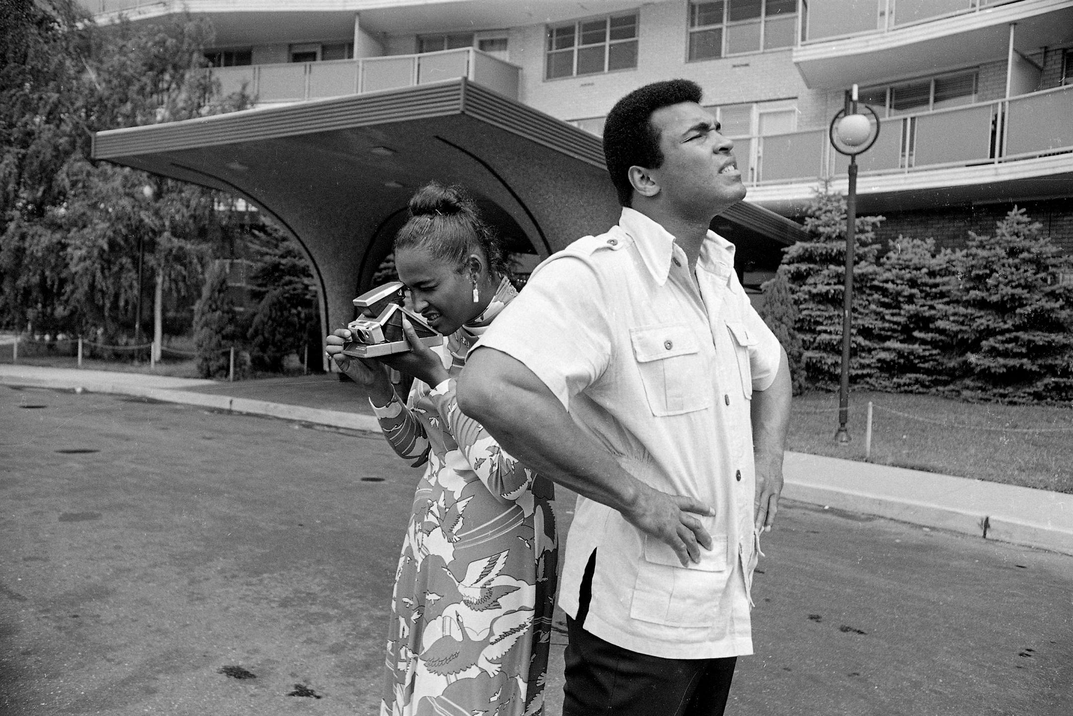 Heavyweight boxing champ Muhammad Ali looks over the front of an apartment building that will temporarily be his new home in Cherry Hill, N.J., while his wife Belinda makes photos in another direction, Aug. 23, 1974. Ali says he is looking for a piece of land. "I'm gonna build me a mansion," he said.