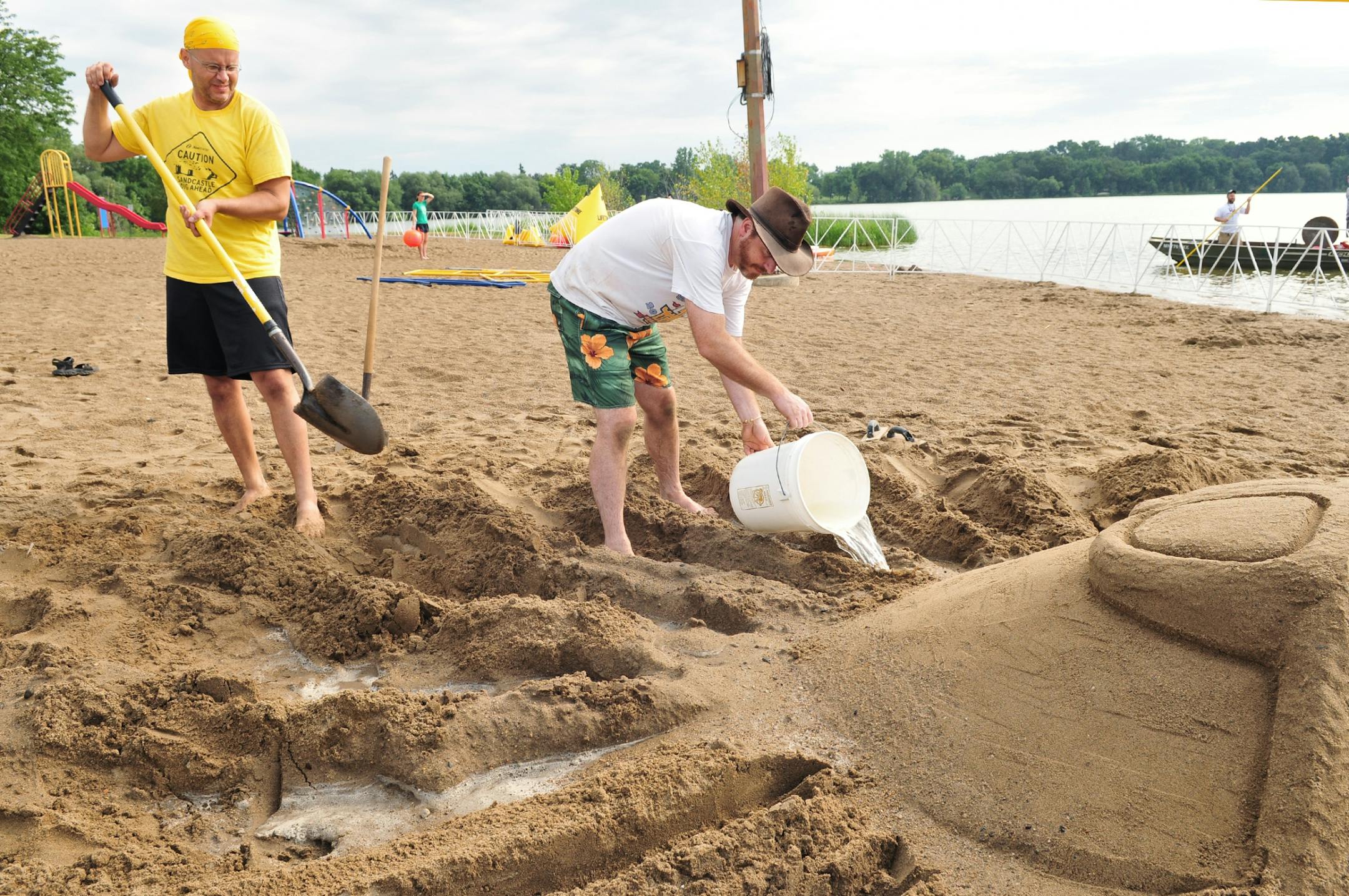 Erik Jorgenson pours water from Lake Nokomis on to an unfinished portion of a sand sculpture to keep it moist enough to continue working. Kendall Bohn uses a shovel to carve the general shape of the sun sculpture's rays.