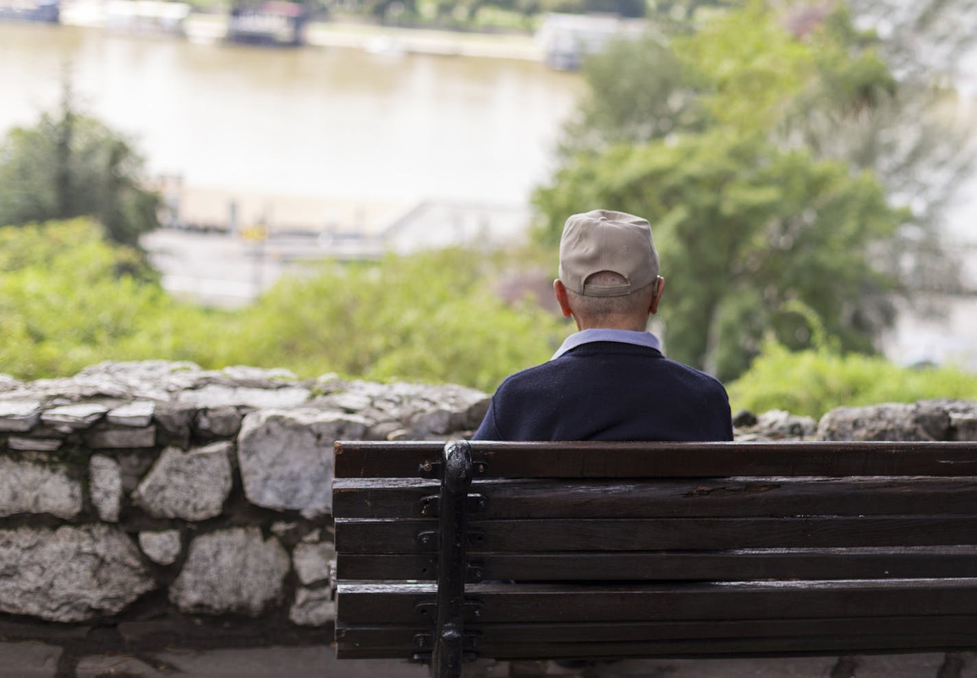 A lonely old man sitting on a bench in a park, looking at river