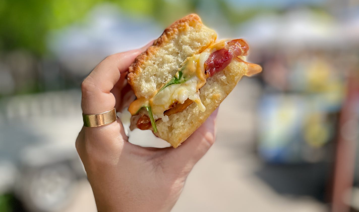 A hand holds up a biscuit sandwiching gooey cheese, crispy prosciutto, fried egg and arugula.