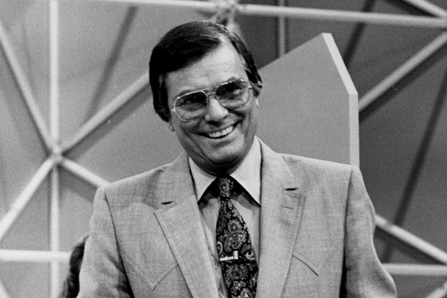 Black and white photo of Peter Marshall in suit and shirt with paisley TV and large wire-framed glasses on game show set.