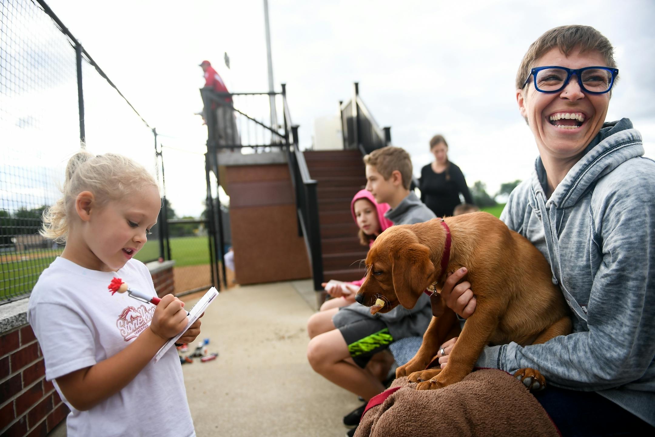 Clara Stalboerger, 5, took a food order from Emily Jonas, of Cold Spring, as she held her 3-year old dog, Arlo, in her lap during Farming Invitational Tournament on Friday, June 23, 2017 in Farming, Minn. Stalboerger's father, Teddy Fleming, plays baseball for the Sartell Stone Poneys, a team in the Sauk Valley League and Jonas is a member of the Schleper family, which Farming's baseball field is named after.