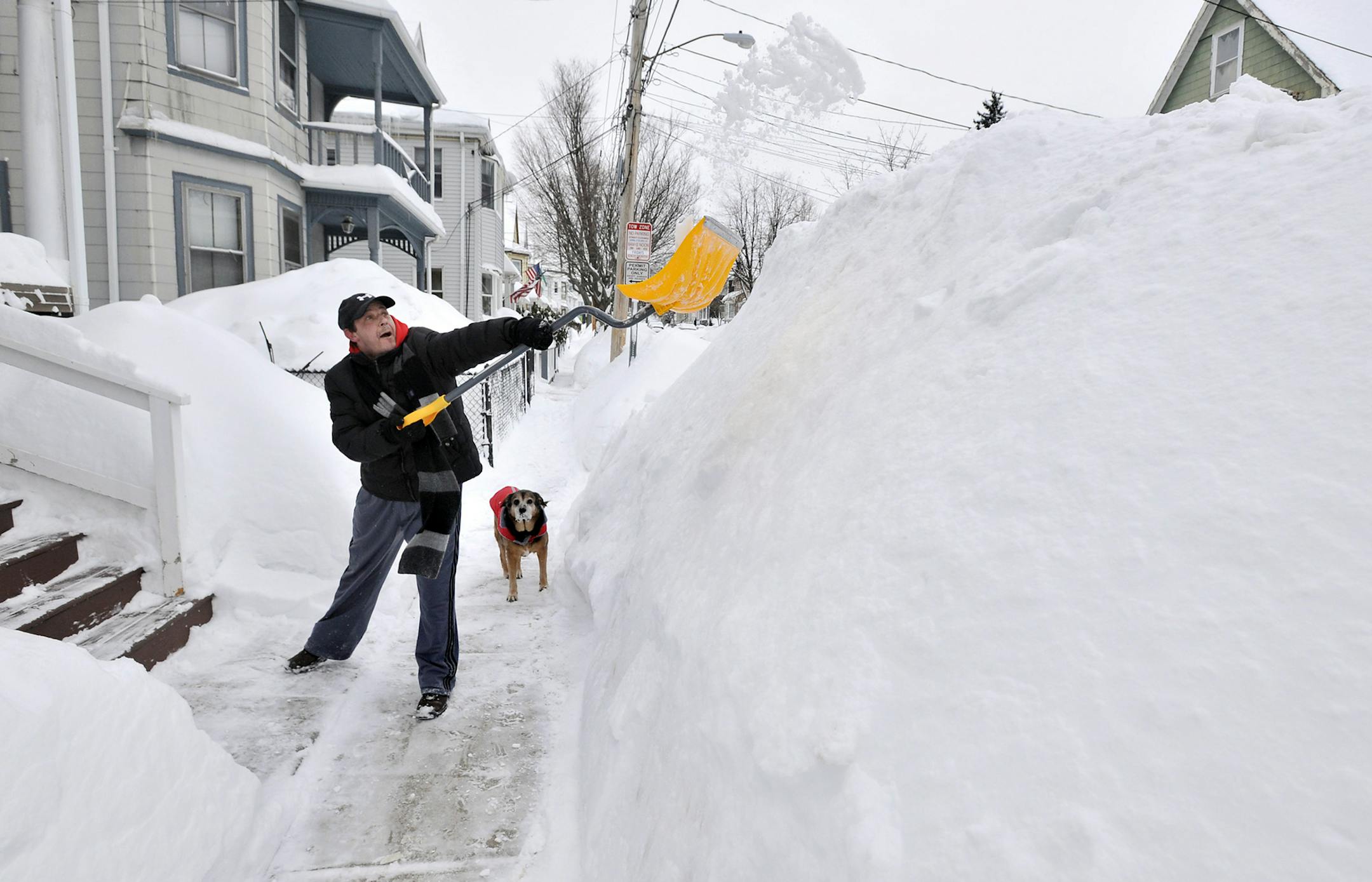 Lee Anderson adds to the pile of snow beside the sidewalk in front of his house in Somerville, Mass., Tuesday, Feb. 10, 2015, as his dog Ace looks on. The latest snowstorm left the Boston area with another two feet of snow and forced the MBTA to suspend all rail service for the day. (AP Photo/Josh Reynolds)
