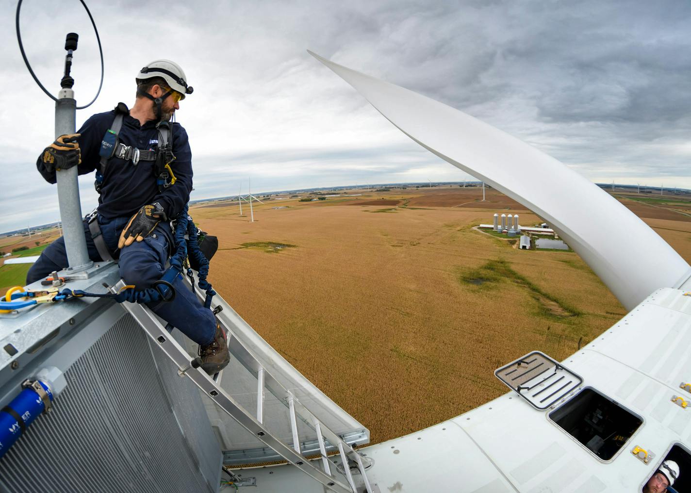 Wind technician Will Osborn perched outside a wind turbine's nacelle, a small room housing the gear box. He and Chris Berg, lower right, were 24 stories up servicing a Vestas wind turbine at the Black Oak wind farm, below, in Sauk Centre, Minn.