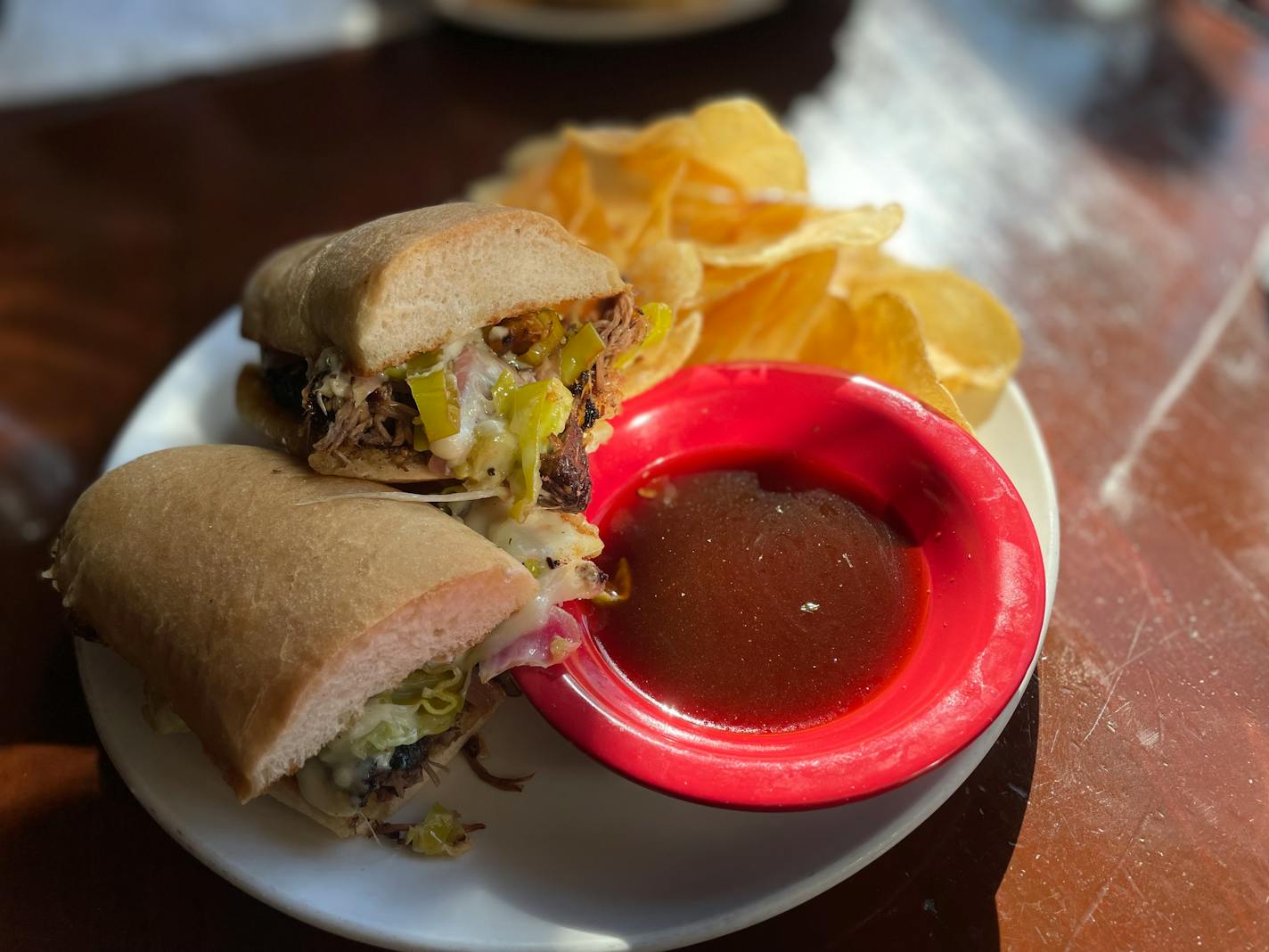 Golden hour light hits a sandwich that's overflowing with banana peppers, braised meat and cheese next to a bowl of jus and a pile of homemade potato chips.