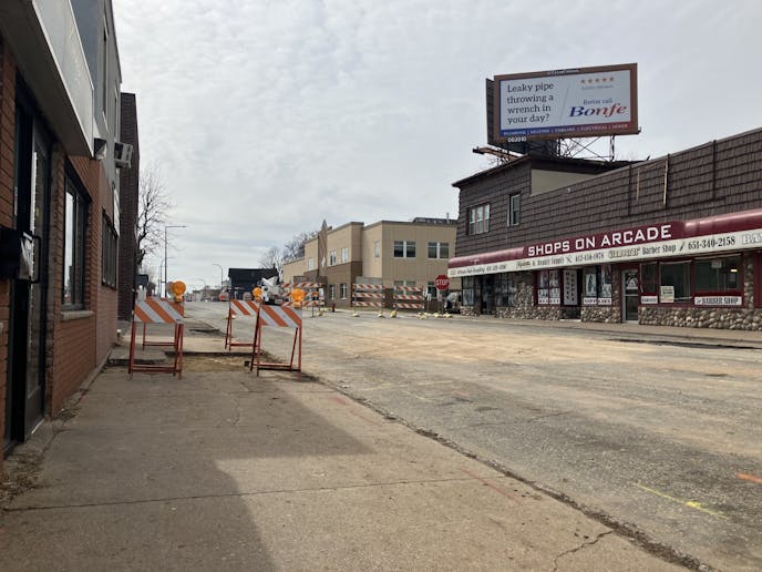 A closed street with orange construction barriers on the sidewalk and at an intersection. A maroon awning on a building reads "Shops on Arcade."