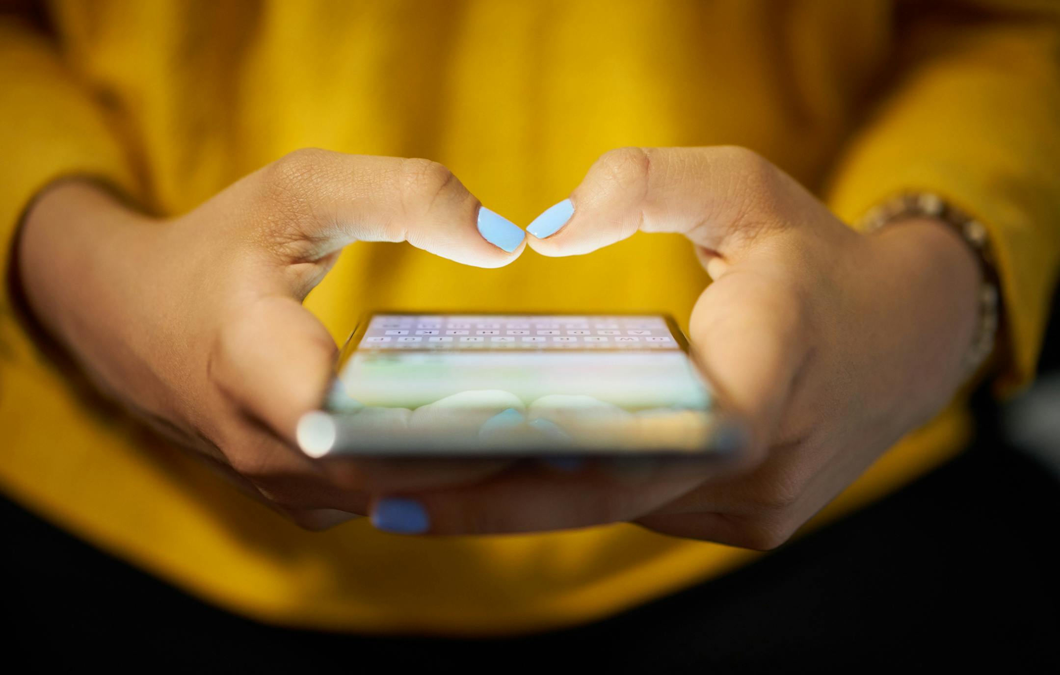 Young woman using cell phone to send text message on social network at night. Closeup of hands with computer laptop in background