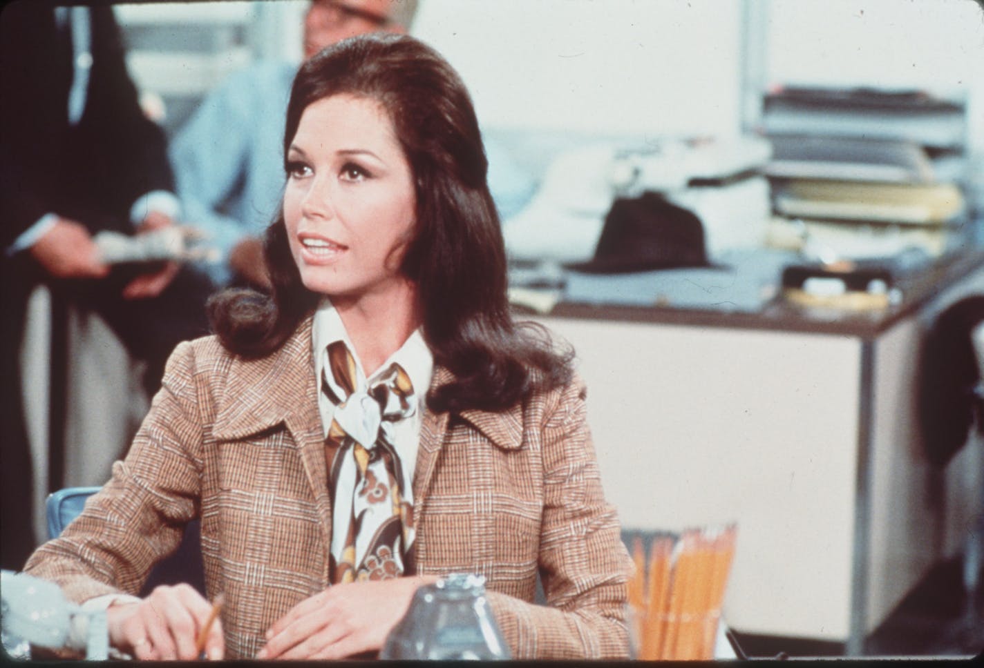 photo of Mary Tyler Moore, seated at a desk in character as Mary Richards