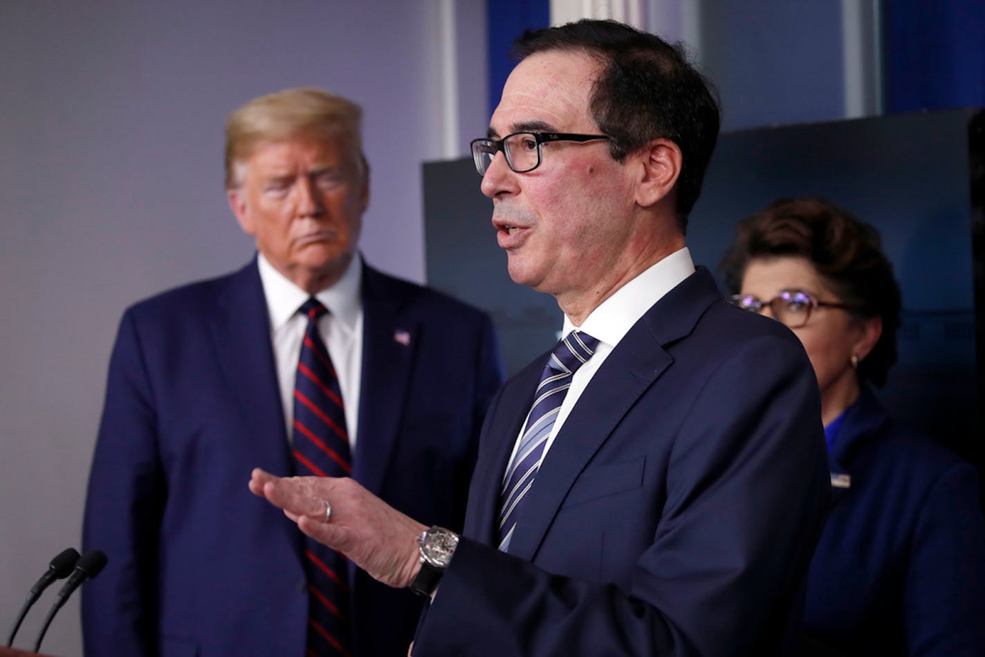 In this April 2, 2020, photo, President Donald Trump and Jovita Carranza, administrator of the Small Business Administration, listen as Treasury Secretary Steven Mnuchin discusses the financial impact of the coronavirus pandemic from the White House in Washington.