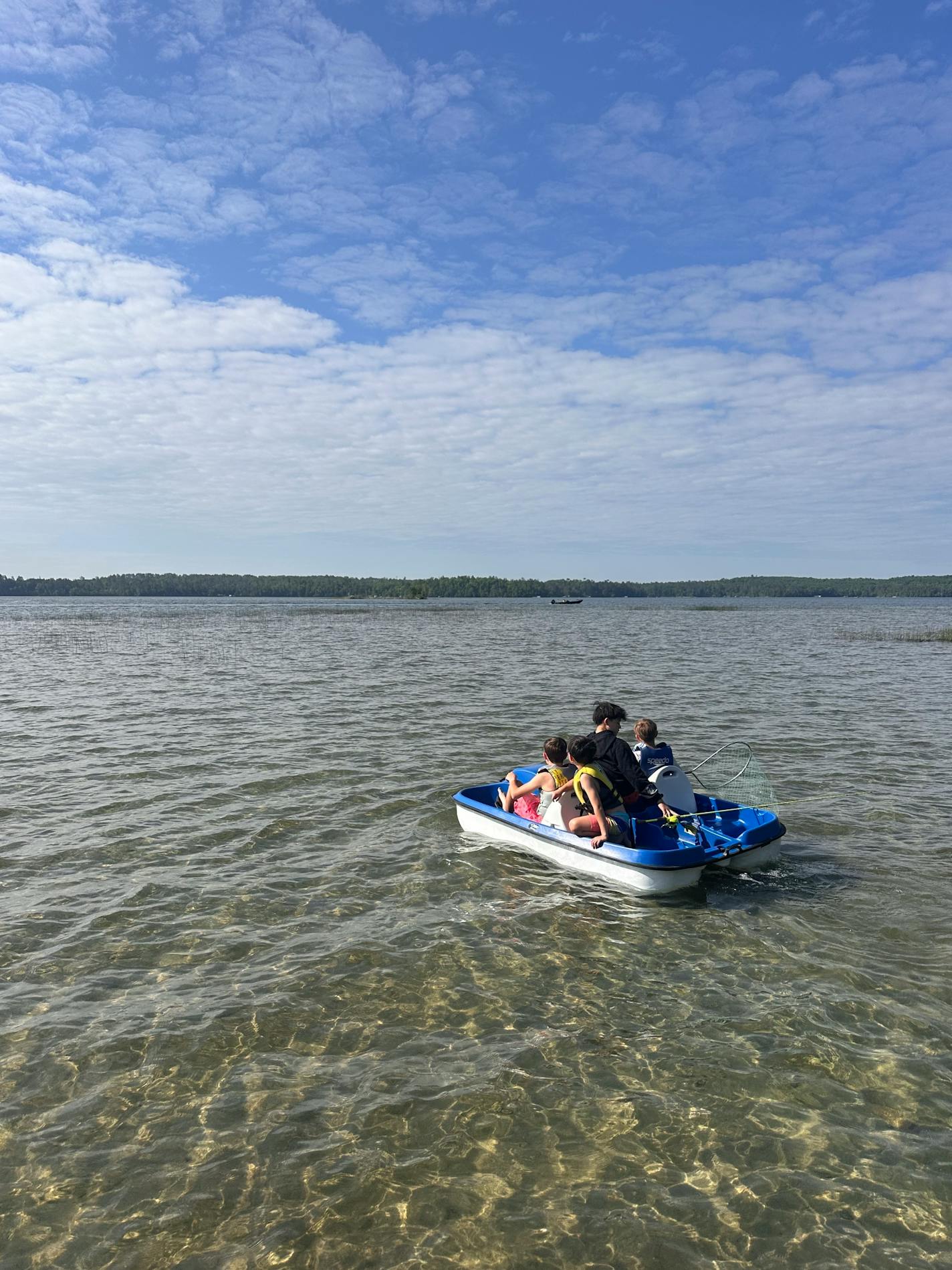 Four young boys wearing life jackets and hoisting fishing poles venture out on a pedal boat in a clear glistening lake with a sandy bottom.