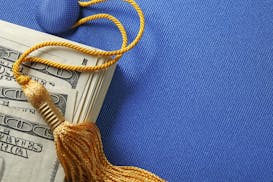 A stack of one hundred dollar bills on top of a blue graduation cap. A gold graduation tassel is draped over the stack of money. The ledft side of ima