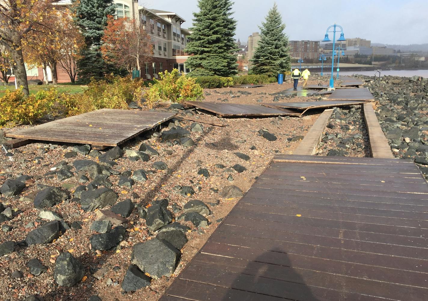 Storm damage to Duluth's Lakewalk in Canal Park, with dislodged boardwalk and debris covering the asphalt path running parallel to it.