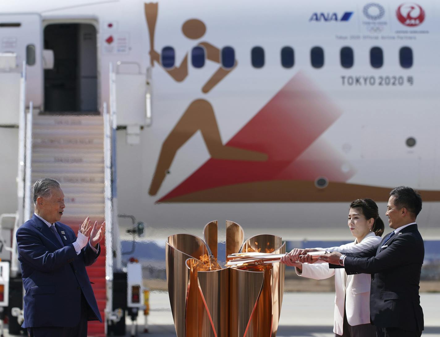 Three-time Olympic gold medalists Tadahiro Nomura, right, and Saori Yoshida light the torch as Tokyo 2020 Olympics chief Yoshiro Mori, left, watches during Olympic Flame Arrival Ceremony at Japan Air Self-Defense Force Matsushima Base in Higashimatsushima in Miyagi Prefecture, north of Tokyo, Friday, March 20, 2020. The Olympic flame from Greece arrived in Japan even as the opening of the the Tokyo Games in four months is in doubt with more voices suggesting the games should to be postponed or c