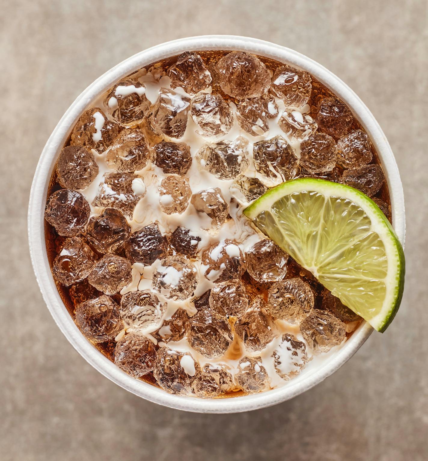 A top-down photo of a cup of brown cola with pebble ice, cream swirled into it and a lime slice on top