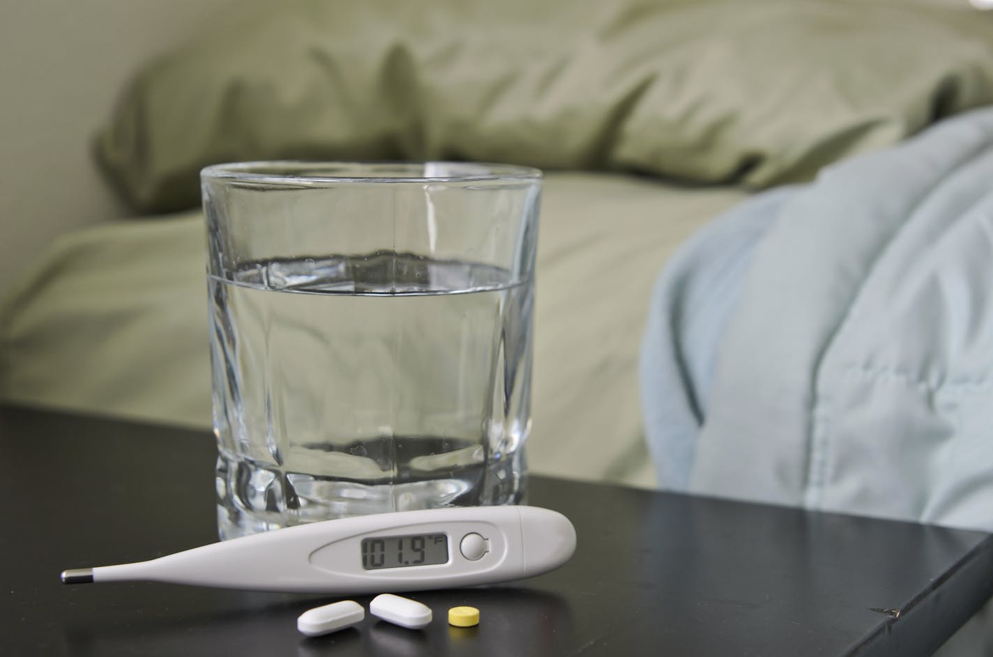 Thermometer showing fever with medicine and cup of water nect to bed