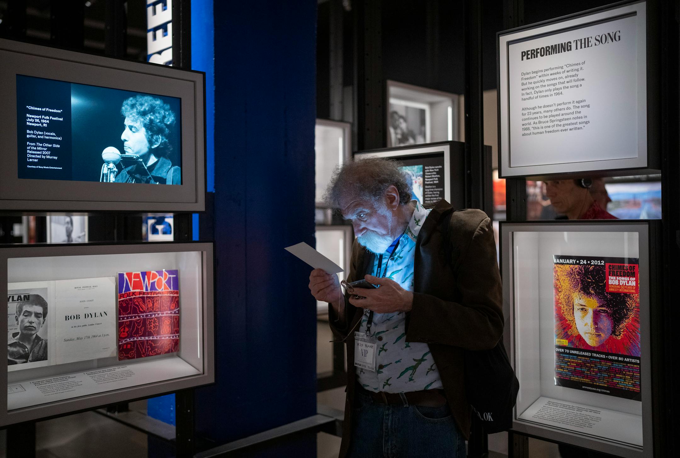 Mitch Blank, a New York collector of Dylanalia and consultant to the Bob Dylan Archive, looked something up on his phone near a display case, left, that contains a program he donated from the 1964 Newport Folk Festival at the Bob Dylan Center in Tulsa, Okla. on Saturday, May 7, 2022. Also in the same case is a program from Dylan's first public London performance in 1964, donated by avid Minnesota collector, Bill Pagel.