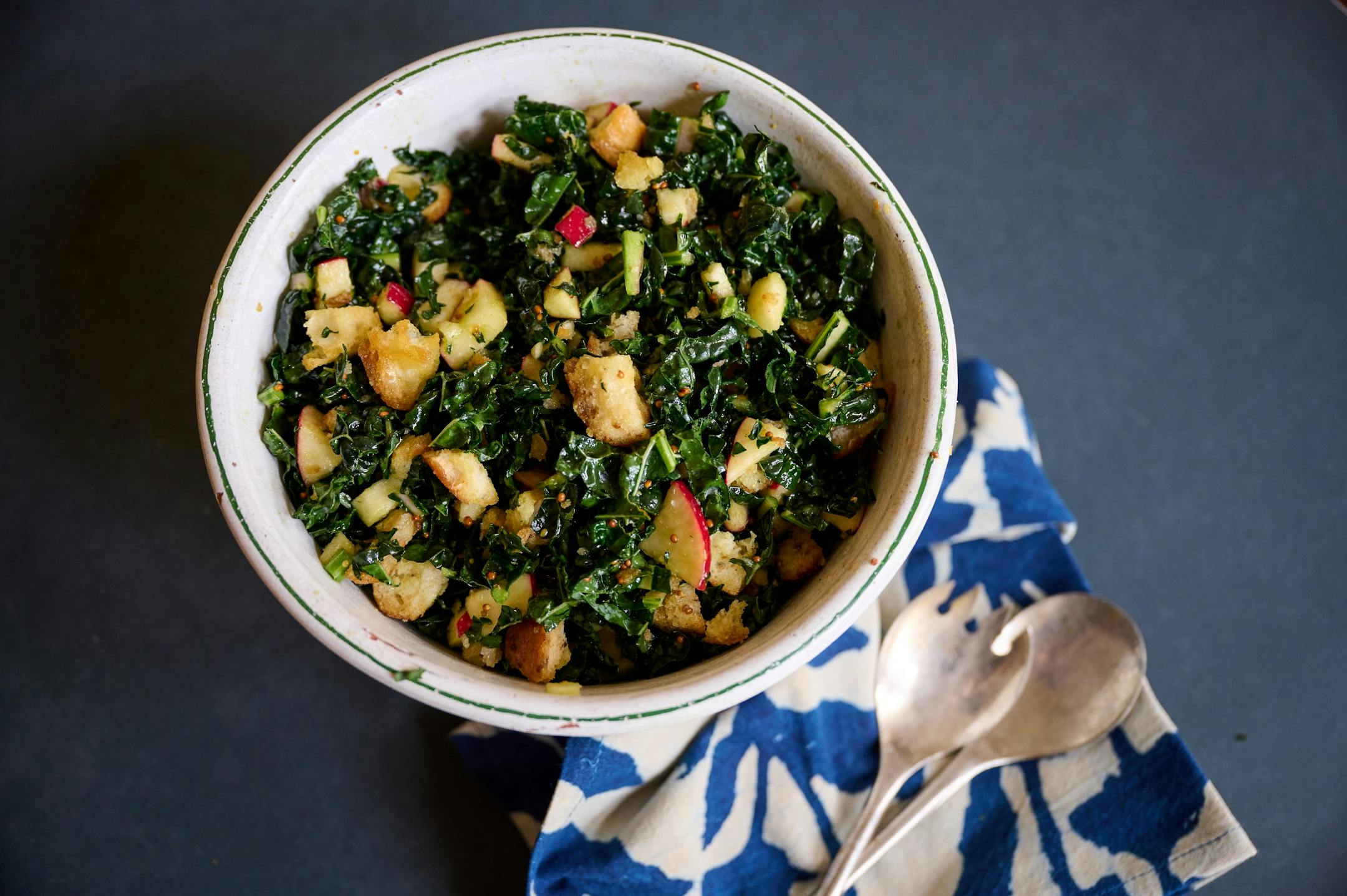 A bowl of kale salad with turkey, stuffing croutons, apples and more alongside a bright blue and white napkin and serving utensils.