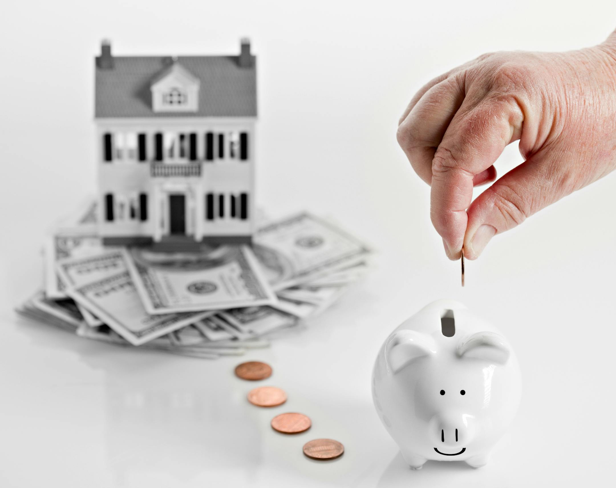 A conceptual image showing a reverse mortgage with a home sitting on a pile of money and a hand dropping coins slowly in a piggy bank. istock photo