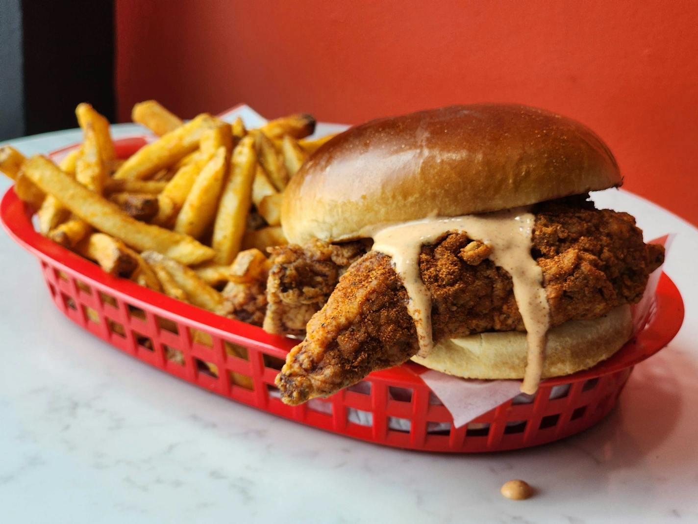 A red basket that contains a sandwich with fried chicken tenders poking out of the shiny bun and pink sauce dripping off of it, and a handful of golden fries