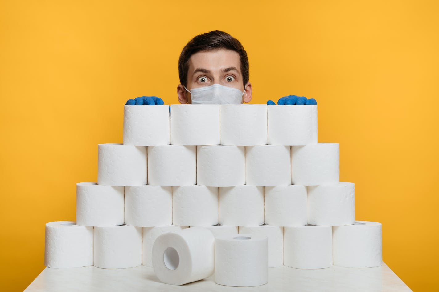 Scared man in a protective face mask is hiding behind toilet paper to protect himself from Coronavirus. Isolated on yellow. Deficit of toilet paper due COVID-19 pandemic, home isolation and quarantine