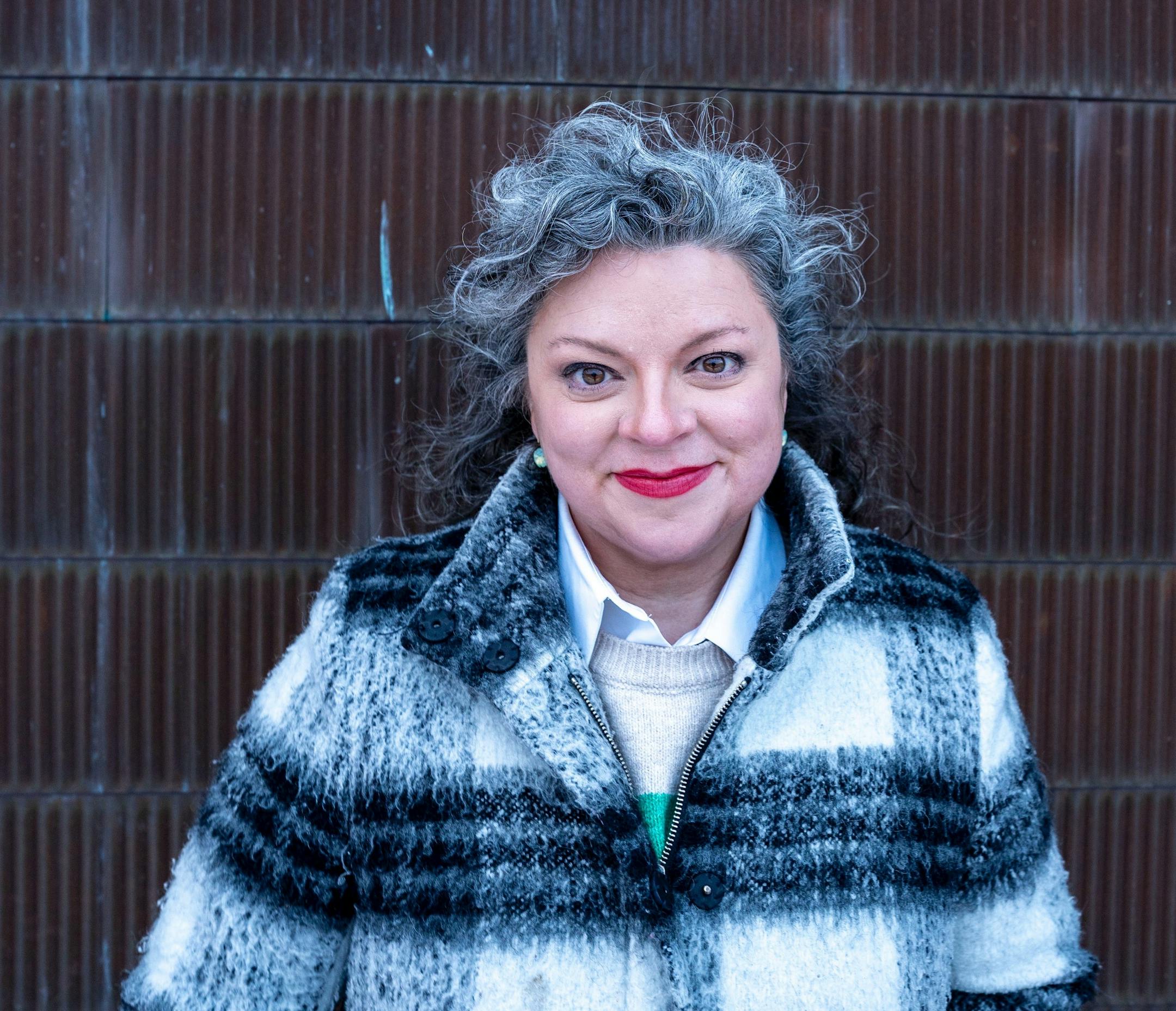 photo of author Kelly Foster Lundquist, in a plaid coat, in front of a brown brick wall