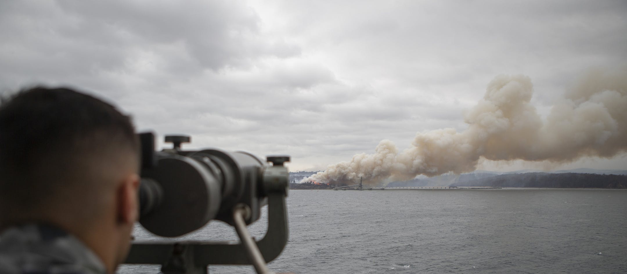 In this photo provided by the Australian Department of Defence on Jan. 6, 2020, Seaman Boatswains Mate Malik El-Leissy watches a burning fire from HMAS Adelaide as the ship arrives at Eden to assist with wildfires. The wildfires have so far scorched an area twice the size of the U.S. state of Maryland. They have destroyed about 2,000 homes. (Able Seaman Thomas Sawtell/ADF via AP)