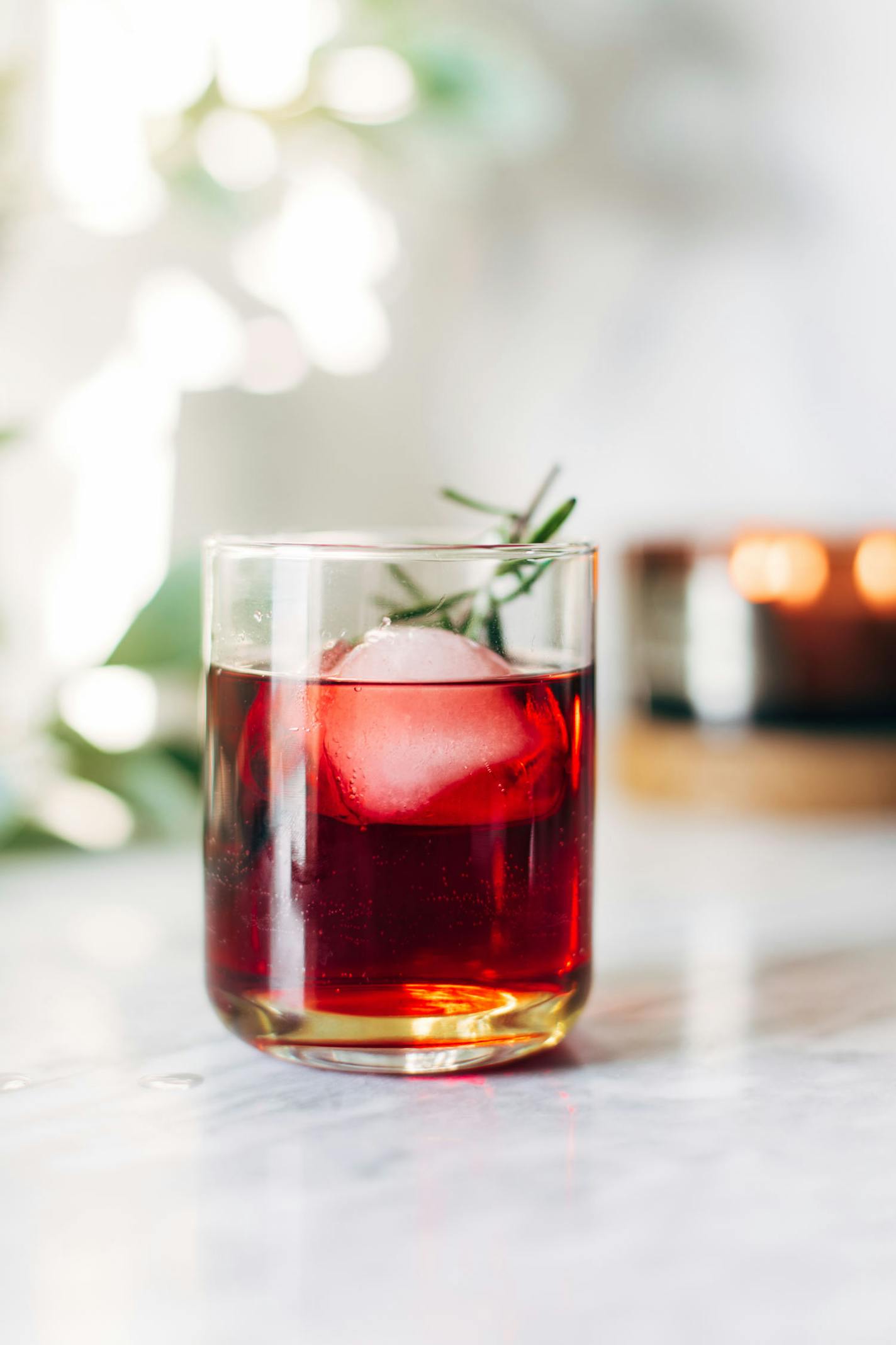 A short cocktail glass of dark red liquid and a sprig of rosemary sits on a white marble countertop.