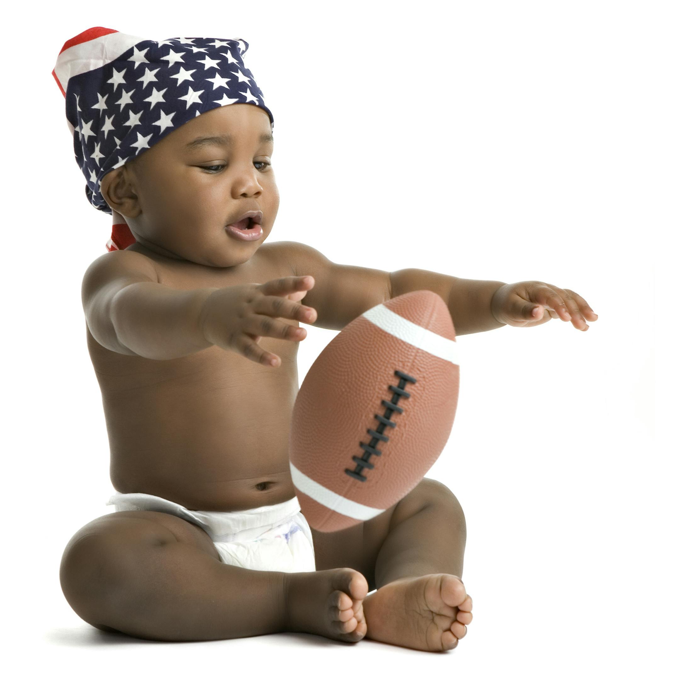 An adorable black baby in a diaper and US flag bandanna playing with a football. Isolated on white.