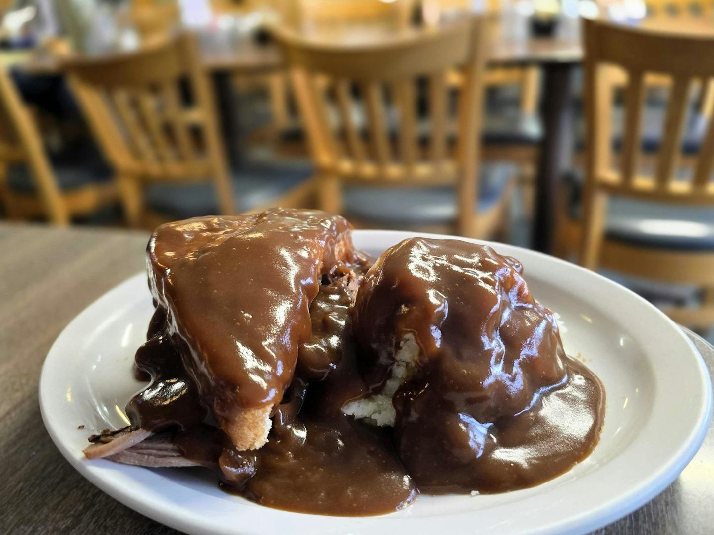 A white plate with a thick brown gravy covering a half beef sandwich and a scoop  of mashed potatoes.