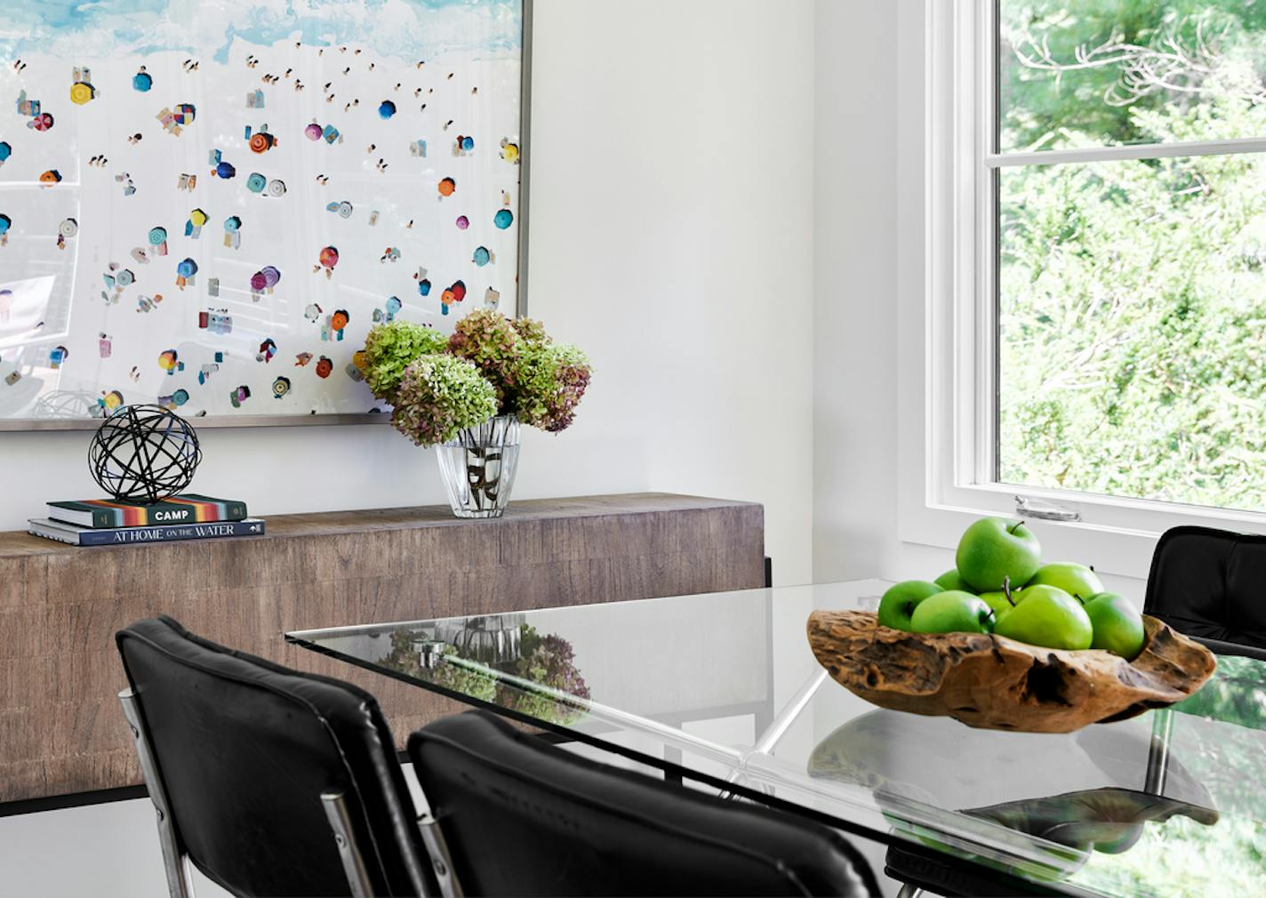 Dining room with a vase of fresh flowers on a buffet that has a colorful modern artwork hanging over it at left and a window with greenery outside to the right. There's a glass table with black leather chairs, with a wooden bowl filled with green apples on top of the table.