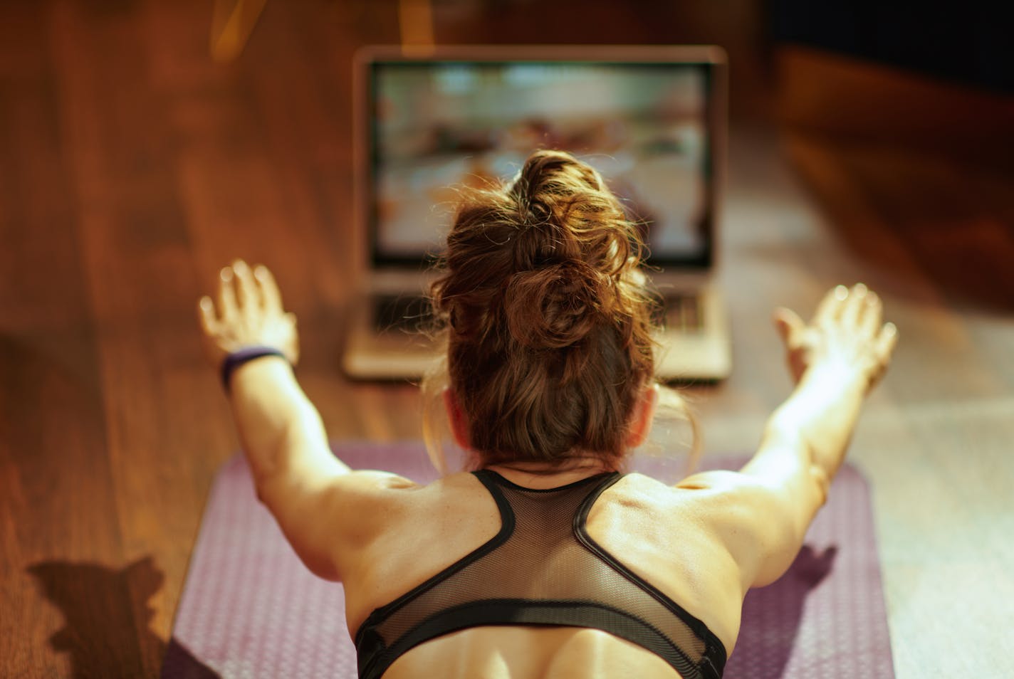 Seen from behind healthy woman in fitness clothes at modern home watching fitness tutorial on internet via laptop while doing exercises on fitness mat.