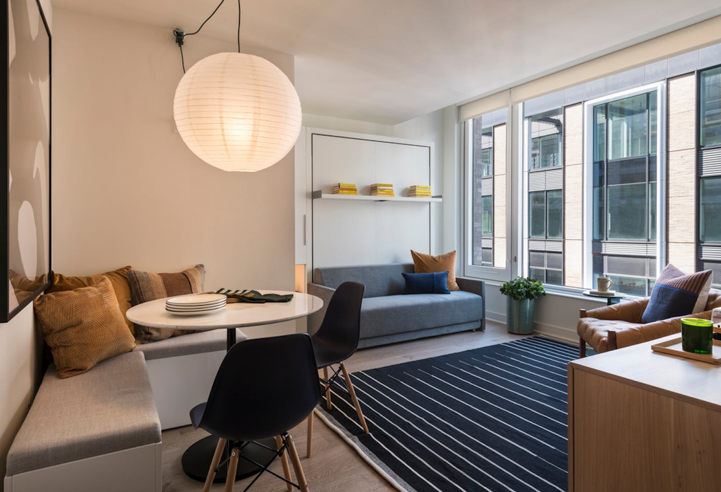 Studio apartment with bench, small white round table and two navy blue chairs at left, with a dark striped rug to the right and a fold-down bed to the back right, with windows to the right showing another building.