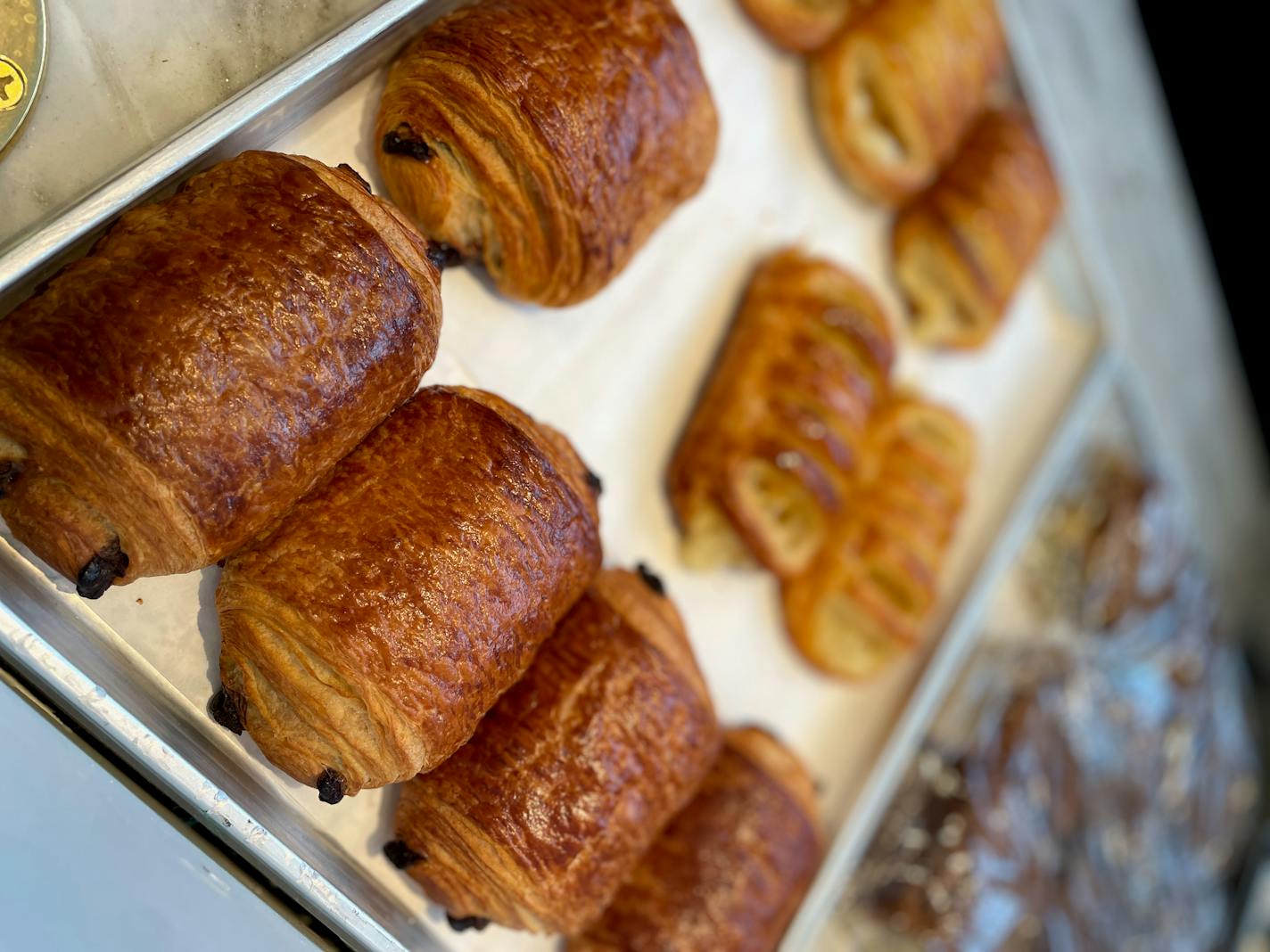 A half sheet pan lined with chocolate croissants with some folded over Danish visible in the background.