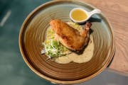 A brown and green clay dish with a crisp-skinned airline cut chicken breast over a light brown sauce, with a white spoon full of a butter sauce on the