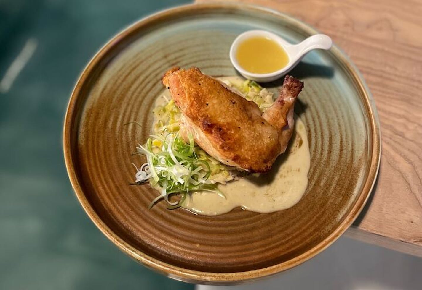 A brown and green clay dish with a crisp-skinned airline cut chicken breast over a light brown sauce, with a white spoon full of a butter sauce on the side.