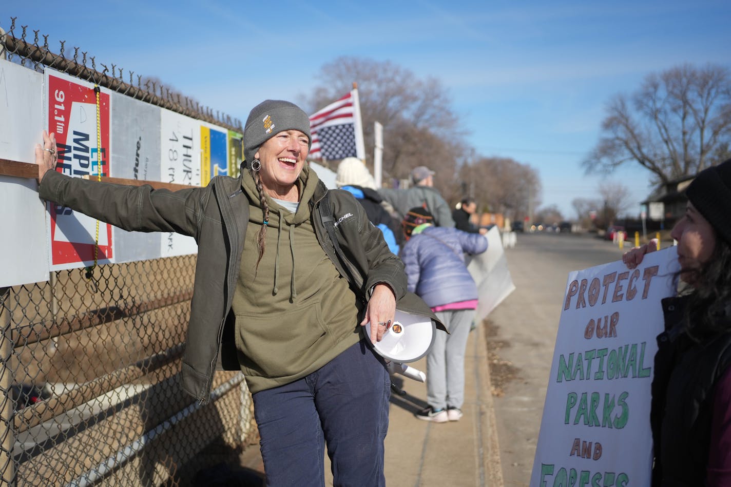 A smiling woman with a megaphone holds up a protest sign about the U.S. Postal Service while talking with another woman holding a sign about protecting national parks.