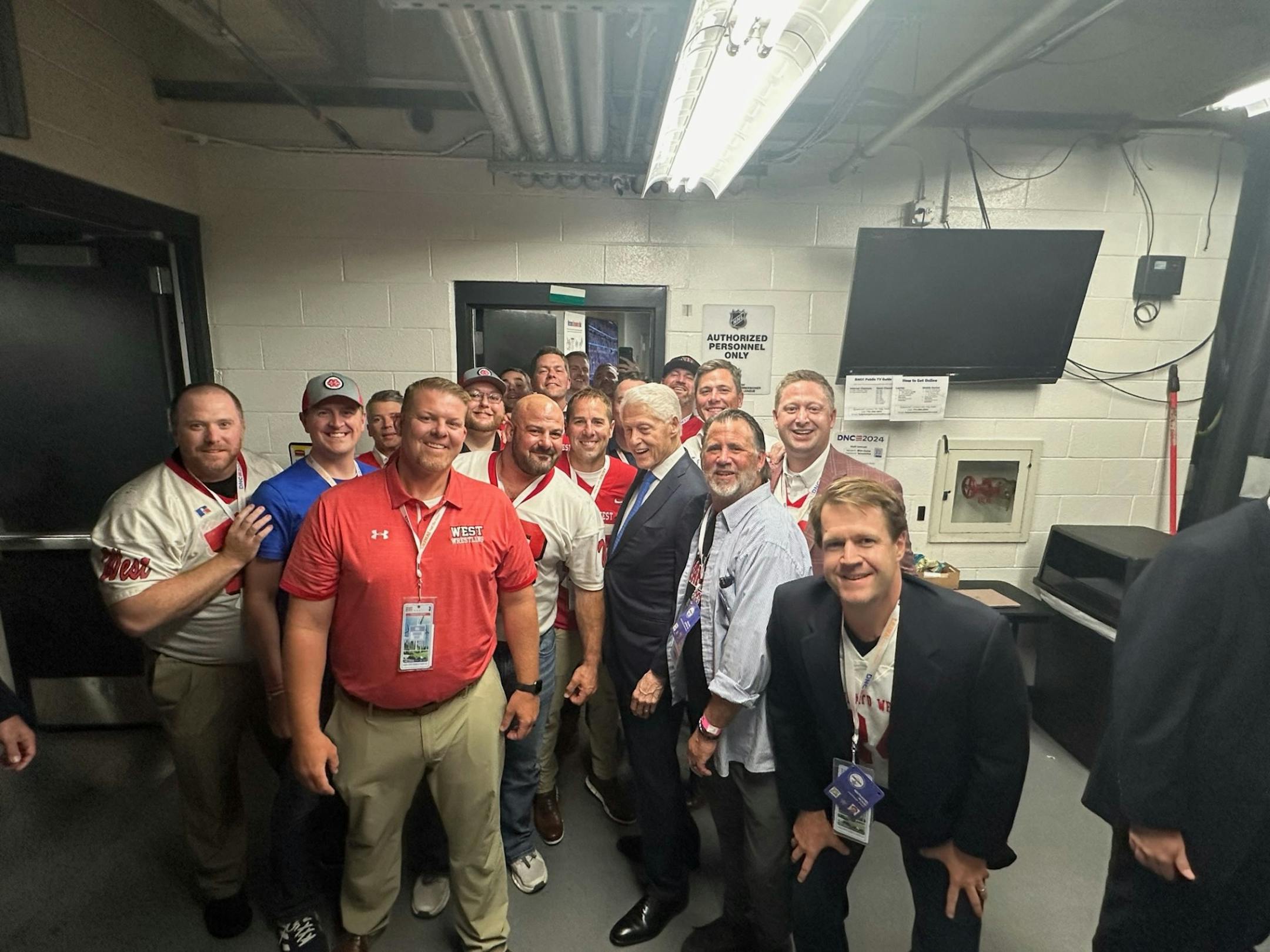Former president Bill Clinton poses with a photo with a crowd of former football players in their late 30s to early 40s. The former football players came to support Gov. Tim Walz, who once coached them, as he runs for vice president.