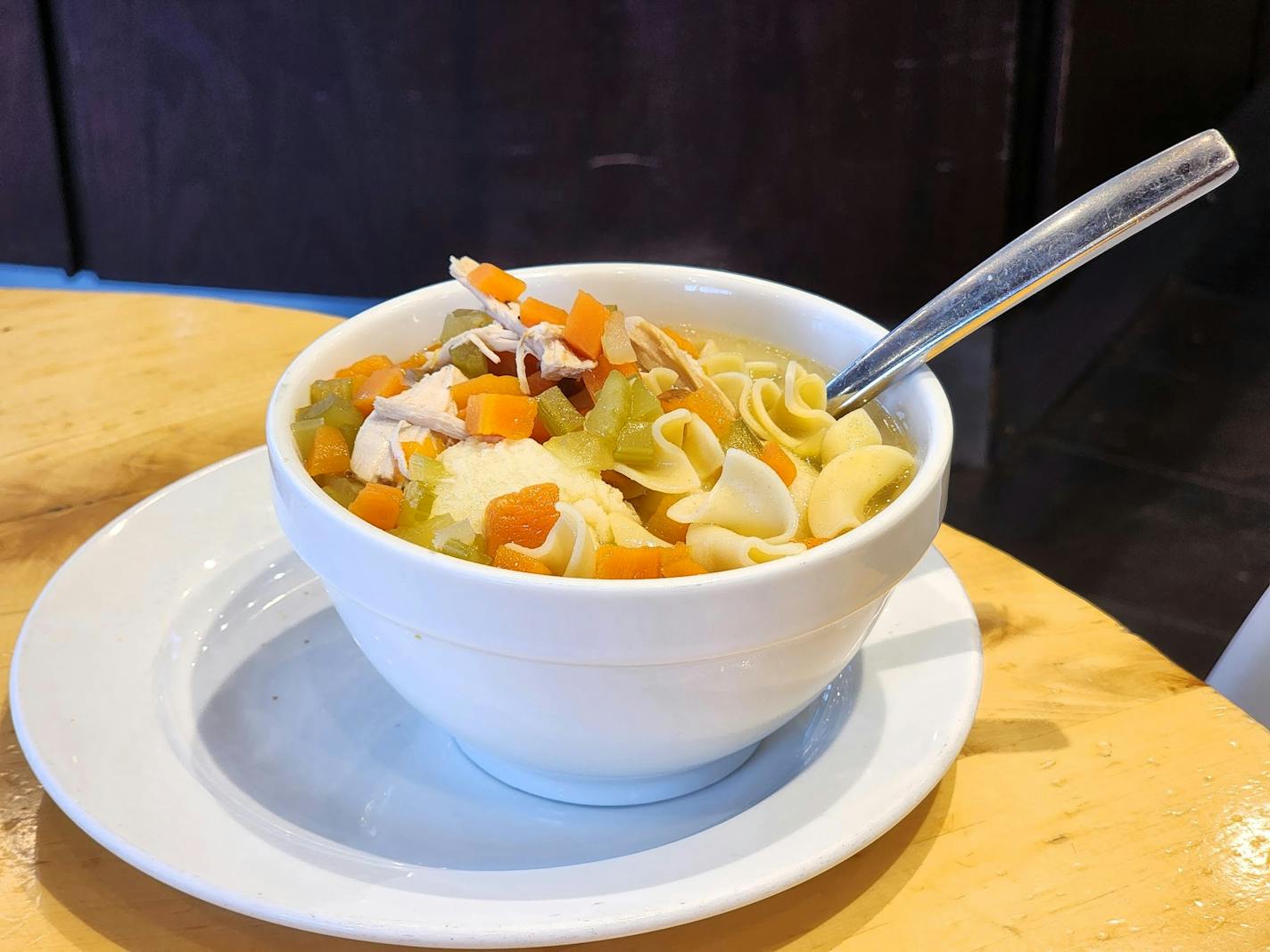 A white bowl on a table filled to the brim with noodles, celery and carrots, and fluffy matzo balls.