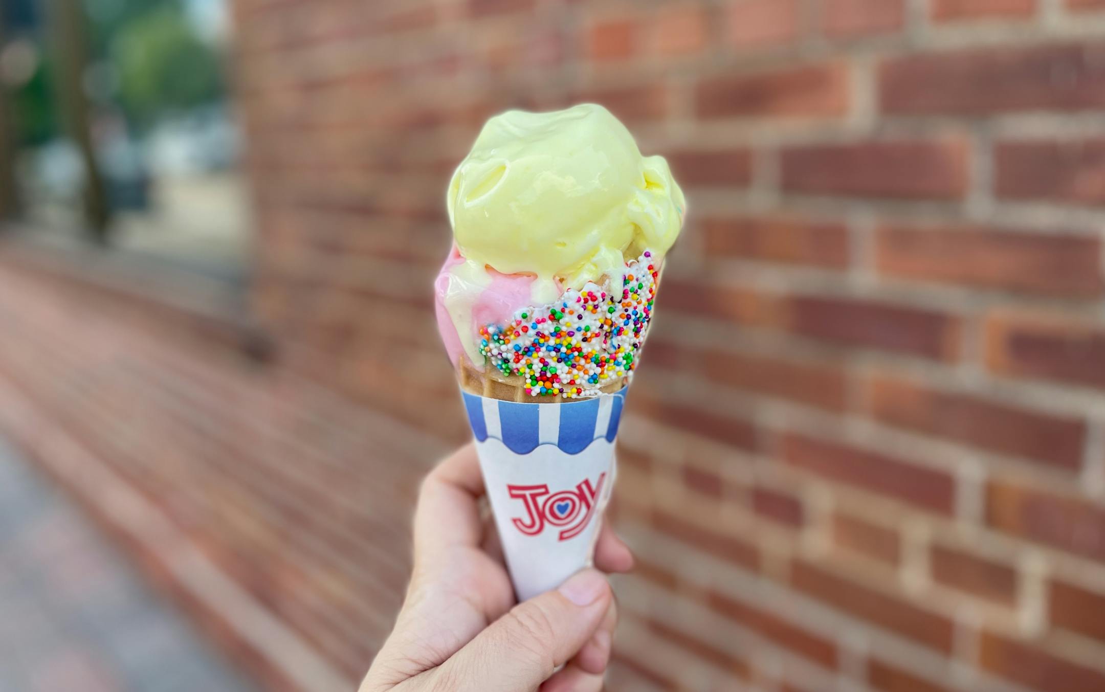 A bright yellow scoop of sorbretto melts into a pink scoop, both atop a confetti sprinkle garnished ice cream cone.